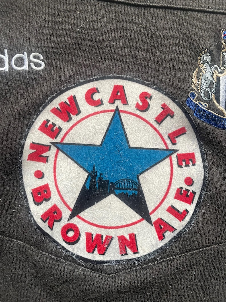 May include: A white and red circular patch with the text "NEWCASTLE BROWN ALE" around a blue five-pointed star with a cityscape inside. The star is on a white background.