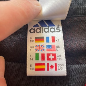 May include: A white Adidas label featuring the Adidas logo and various country flags. The label includes size details and country codes like GB and CDN L/G. The label is being held by a hand.