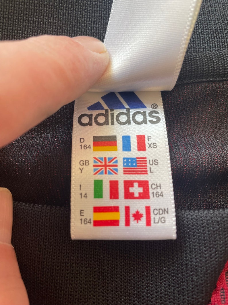 May include: A white Adidas label featuring the Adidas logo and various country flags. The label includes size details and country codes like GB and CDN L/G. The label is being held by a hand.
