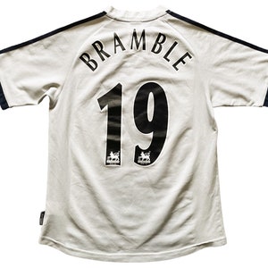 Puede incluir: Camiseta de f&uacute;tbol blanca con ribetes negros y azul marino. La parte posterior de la camiseta tiene el nombre "BRAMBLE" y el n&uacute;mero "19" impresos en negro.