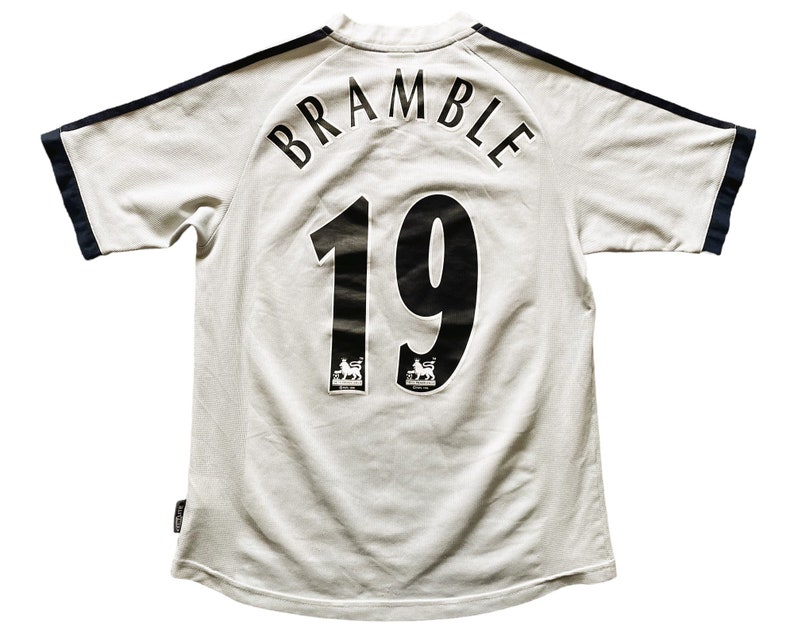 Puede incluir: Camiseta de f&uacute;tbol blanca con ribetes negros y azul marino. La parte posterior de la camiseta tiene el nombre "BRAMBLE" y el n&uacute;mero "19" impresos en negro.