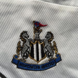 May include: White embroidered patch with a black and white shield, two gold seahorses, and a red and white flag. The text "Newcastle United" is on a blue ribbon.