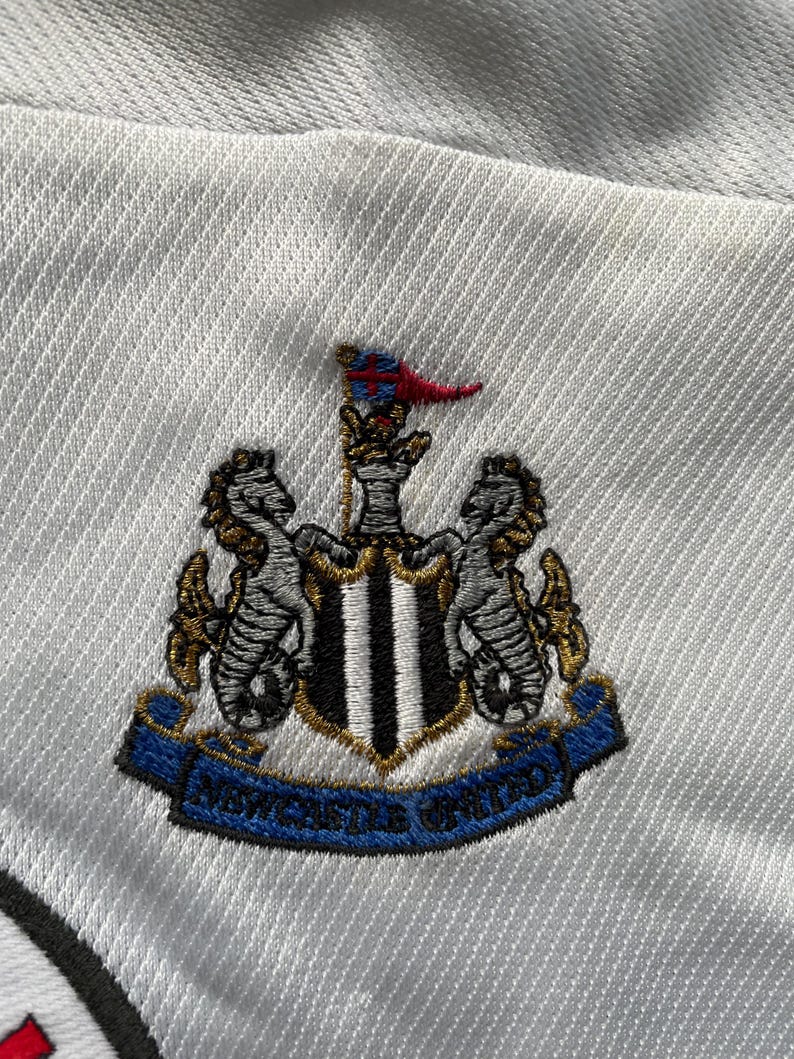 May include: White embroidered patch with a black and white shield, two gold seahorses, and a red and white flag. The text "Newcastle United" is on a blue ribbon.
