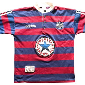 May include: A retro Newcastle United football shirt, featuring red and navy blue stripes. The Adidas logo is present, alongside the club crest and a Newcastle Brown Ale badge. The shirt has a white collar and short sleeves.