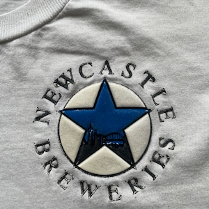 May include: White t-shirt with a black and white embroidered logo that says "Newcastle Breweries". The logo features a blue five-pointed star with a cityscape inside.