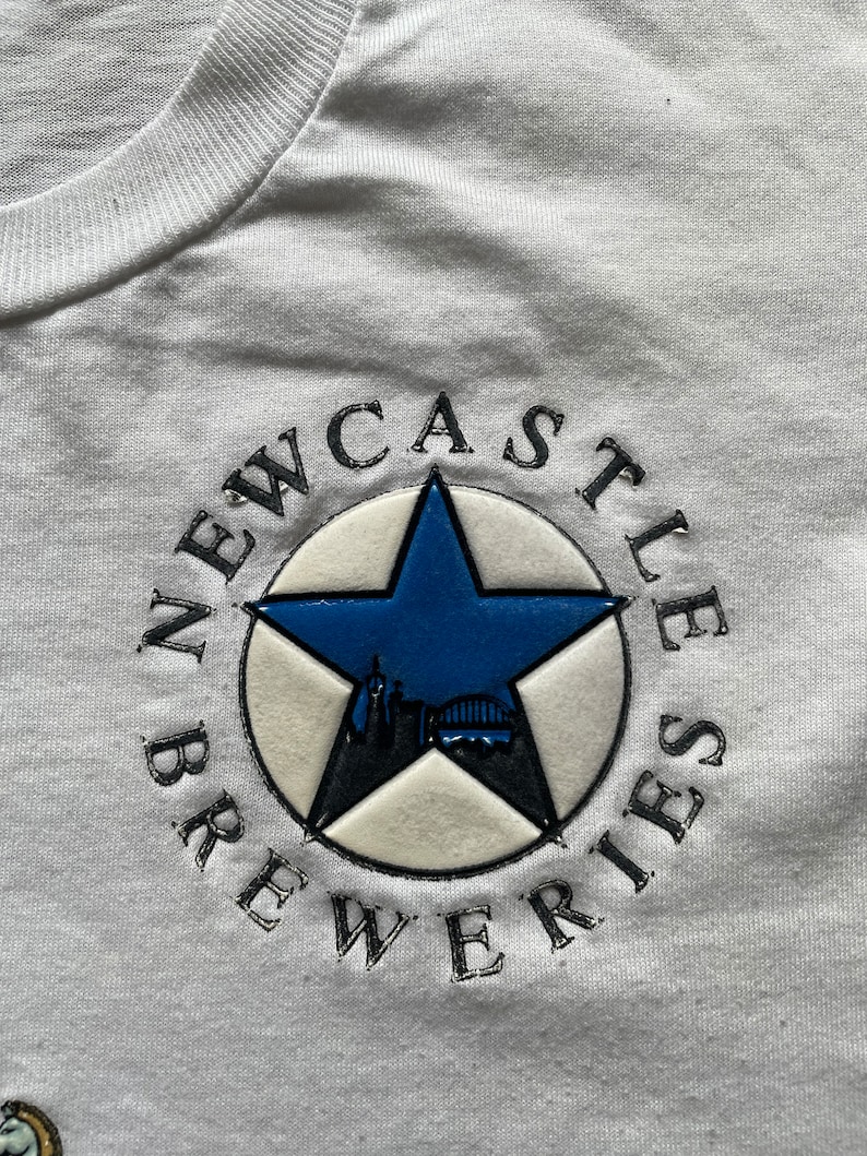 May include: White t-shirt with a black and white embroidered logo that says "Newcastle Breweries". The logo features a blue five-pointed star with a cityscape inside.