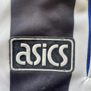 May include: Close-up of a black and white striped athletic jersey with the "asics" logo patch. The logo is white on a black rectangular background. The jersey has a blue stripe.