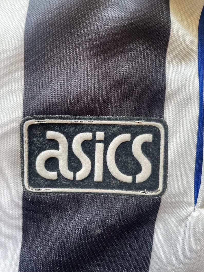 May include: Close-up of a black and white striped athletic jersey with the "asics" logo patch. The logo is white on a black rectangular background. The jersey has a blue stripe.
