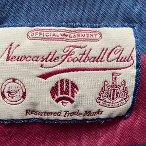 May include: A close-up of a label from a Newcastle Football Club garment. The label is rectangular with a cream background and features the club's name in burgundy script, along with several emblems and the text "Registered Trade Marks."