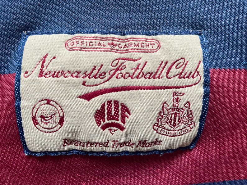 May include: A close-up of a label from a Newcastle Football Club garment. The label is rectangular with a cream background and features the club's name in burgundy script, along with several emblems and the text "Registered Trade Marks."
