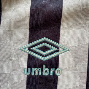 Pode incluir: Uma camisa de futebol listrada branca e preta com um logotipo Umbro azul e branco e a palavra "Umbro" em letras azuis.