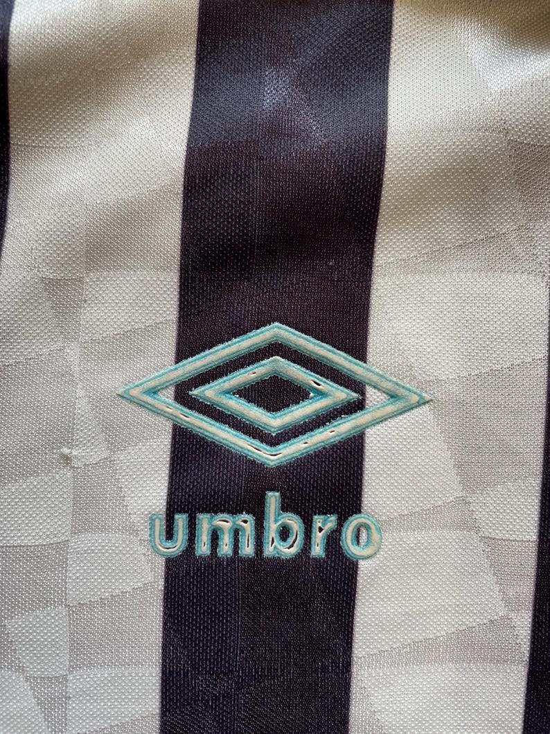 Pode incluir: Uma camisa de futebol listrada branca e preta com um logotipo Umbro azul e branco e a palavra "Umbro" em letras azuis.