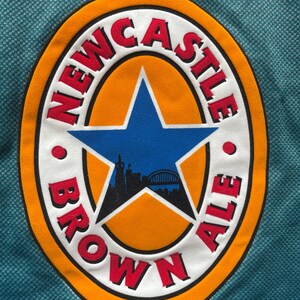 May include: Close-up of a teal fabric with a circular patch. The patch features the words "NEWCASTLE BROWN ALE" in red lettering around a blue star and a silhouette of a city skyline.