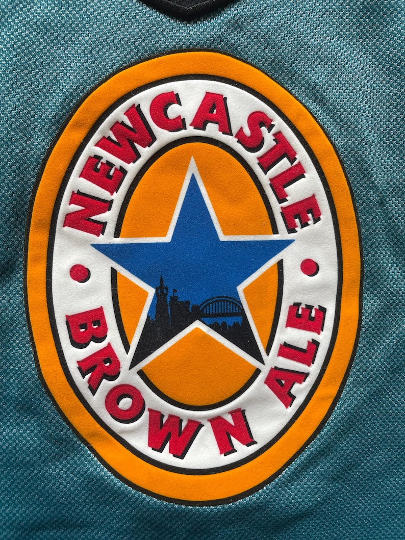 May include: Close-up of a teal fabric with a circular patch. The patch features the words "NEWCASTLE BROWN ALE" in red lettering around a blue star and a silhouette of a city skyline.