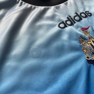 K&ouml;nnte beinhalten: Ein hellblaues T-Shirt mit schwarzem Kragen und dem Schriftzug "adidas" in schwarzer Schrift auf der Vorderseite. Das Shirt zeigt au&szlig;erdem ein Wappen mit einem L&ouml;wen und einer Krone.