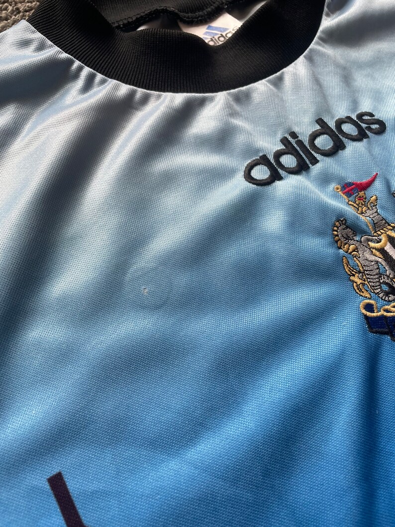 K&ouml;nnte beinhalten: Ein hellblaues T-Shirt mit schwarzem Kragen und dem Schriftzug "adidas" in schwarzer Schrift auf der Vorderseite. Das Shirt zeigt au&szlig;erdem ein Wappen mit einem L&ouml;wen und einer Krone.