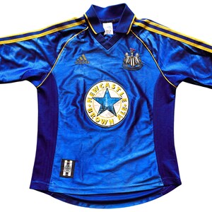 K&ouml;nnte beinhalten: Ein blaues Fu&szlig;balltrikot mit gelben und schwarzen Akzenten. Das Trikot tr&auml;gt das Logo von Newcastle United und die Worte "Newcastle Brown Ale" auf der Vorderseite.