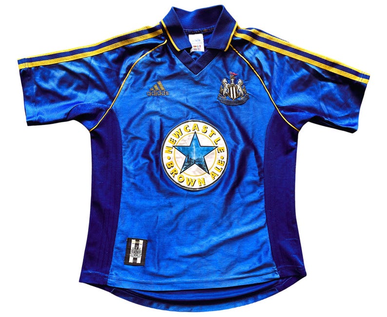 K&ouml;nnte beinhalten: Ein blaues Fu&szlig;balltrikot mit gelben und schwarzen Akzenten. Das Trikot tr&auml;gt das Logo von Newcastle United und die Worte "Newcastle Brown Ale" auf der Vorderseite.