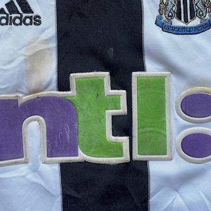 May include: A white and black striped soccer jersey with the Adidas logo on the left chest, the Newcastle United crest on the right chest, and the NTL logo in purple, green, and white across the front.