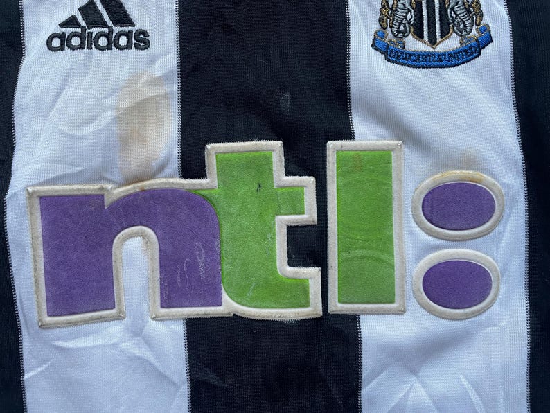 May include: A white and black striped soccer jersey with the Adidas logo on the left chest, the Newcastle United crest on the right chest, and the NTL logo in purple, green, and white across the front.