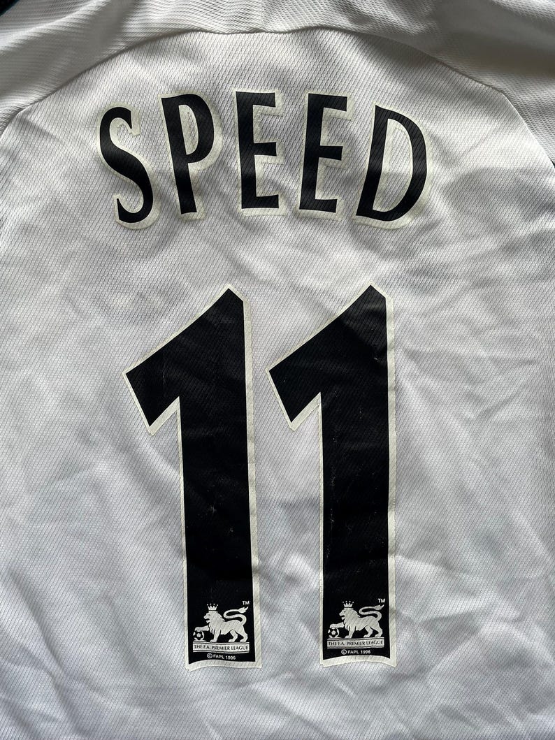 Puede incluir: Camiseta de f&uacute;tbol blanca con el nombre "SPEED" en letras negras grandes. Debajo, dos grandes n&uacute;meros negros "11". El logotipo de la Premier League est&aacute; en la parte inferior de cada n&uacute;mero.
