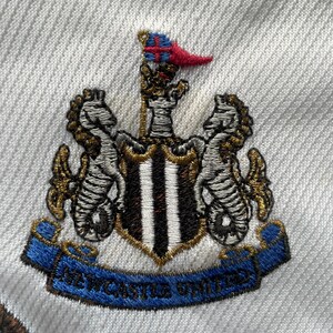 Puede incluir: Escudo bordado del Newcastle United sobre tela blanca. El escudo presenta un escudo con rayas negras y blancas, flanqueado por caballitos de mar y una pancarta azul con la inscripci&oacute;n "NEWCASTLE UNITED".