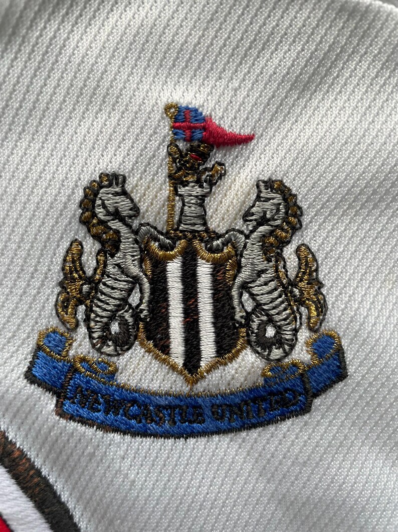 Puede incluir: Escudo bordado del Newcastle United sobre tela blanca. El escudo presenta un escudo con rayas negras y blancas, flanqueado por caballitos de mar y una pancarta azul con la inscripci&oacute;n "NEWCASTLE UNITED".