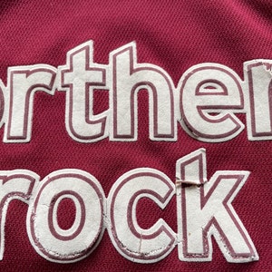 Pu&ograve; includere: Primo piano di una maglietta color granata con la scritta bianca "Northern Rock".