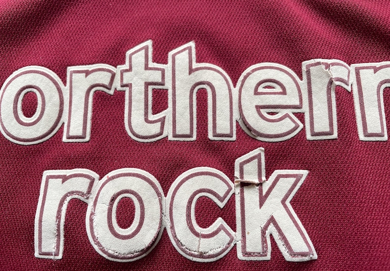 Pu&ograve; includere: Primo piano di una maglietta color granata con la scritta bianca "Northern Rock".