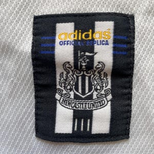 May include: A close-up of a black and white patch with the Adidas logo and the text "OFFICIAL REPLICA" in gold. The patch features the Newcastle United crest and the words "NEWCASTLE UNITED".