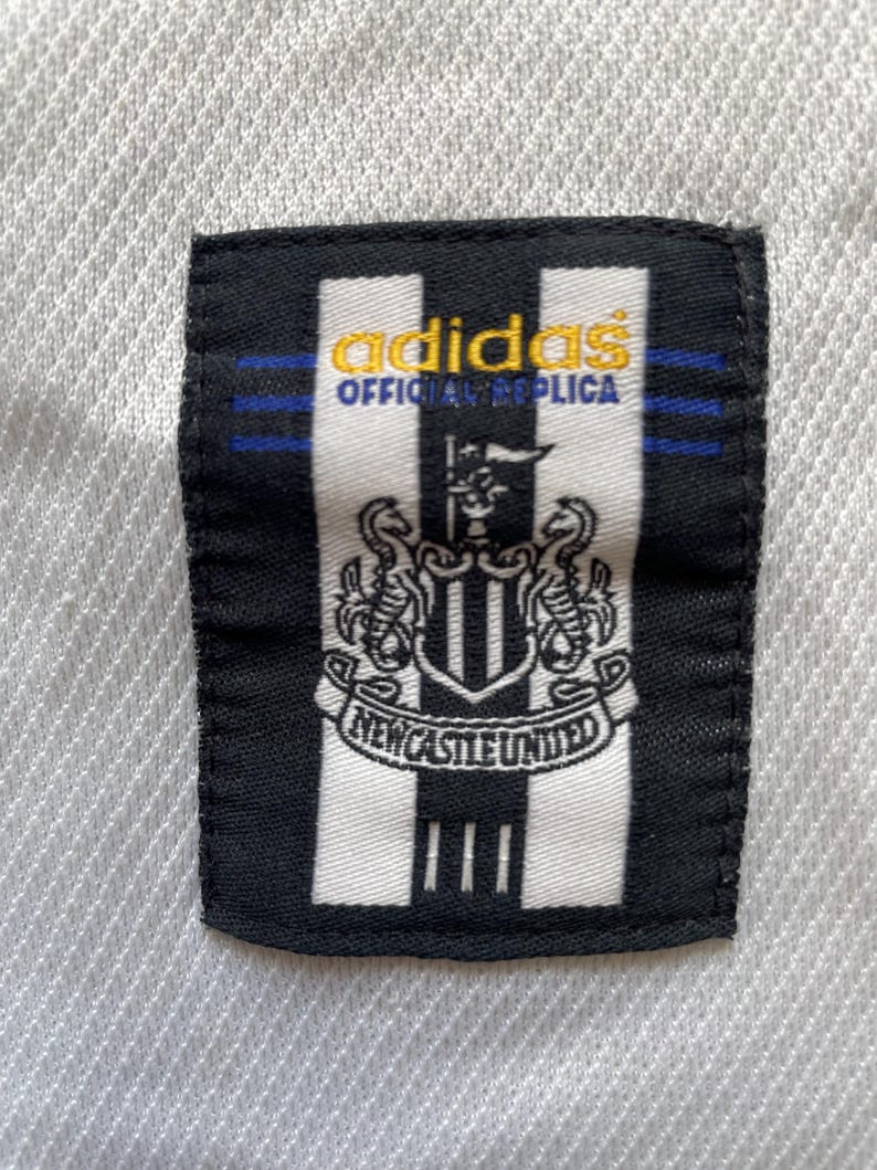 May include: A close-up of a black and white patch with the Adidas logo and the text "OFFICIAL REPLICA" in gold. The patch features the Newcastle United crest and the words "NEWCASTLE UNITED".