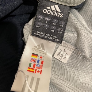 Puede incluir: Una etiqueta Adidas blanca y negra con el texto "adidas" en negro, "MADE IN PORTUGAL" y "FABRIQUE AU PORTUGAL" en negro, e informaci&oacute;n de tallas en negro. La etiqueta est&aacute; unida a una camiseta Adidas gris y negra.