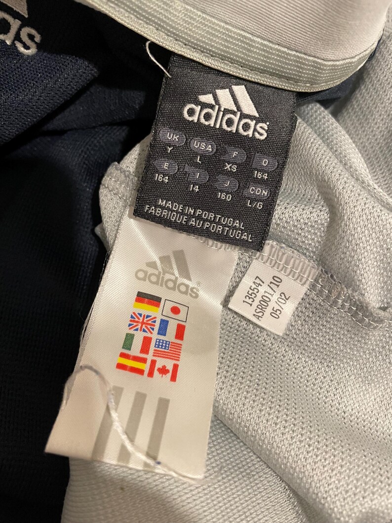 Puede incluir: Una etiqueta Adidas blanca y negra con el texto "adidas" en negro, "MADE IN PORTUGAL" y "FABRIQUE AU PORTUGAL" en negro, e informaci&oacute;n de tallas en negro. La etiqueta est&aacute; unida a una camiseta Adidas gris y negra.