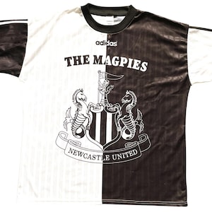 May include: A black and white Newcastle United football shirt. The shirt features the text "THE MAGPIES" and the club crest. The Adidas logo is present. The shirt is split vertically, with one side black and the other white.