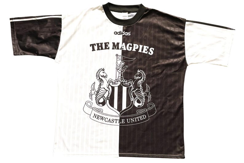 May include: A black and white Newcastle United football shirt. The shirt features the text "THE MAGPIES" and the club crest. The Adidas logo is present. The shirt is split vertically, with one side black and the other white.