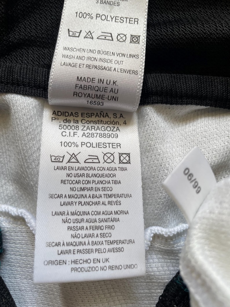 May include: A close-up shot of a white clothing label with black text. The label details care instructions, including washing and ironing guidance. It also states "Made in U.K." and features the Adidas logo and address.