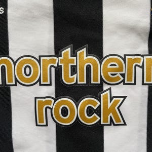May include: Close-up of a black and white striped jersey. The jersey features the text "northern rock" in gold lettering with a black outline. The Adidas logo is visible in the top left corner.