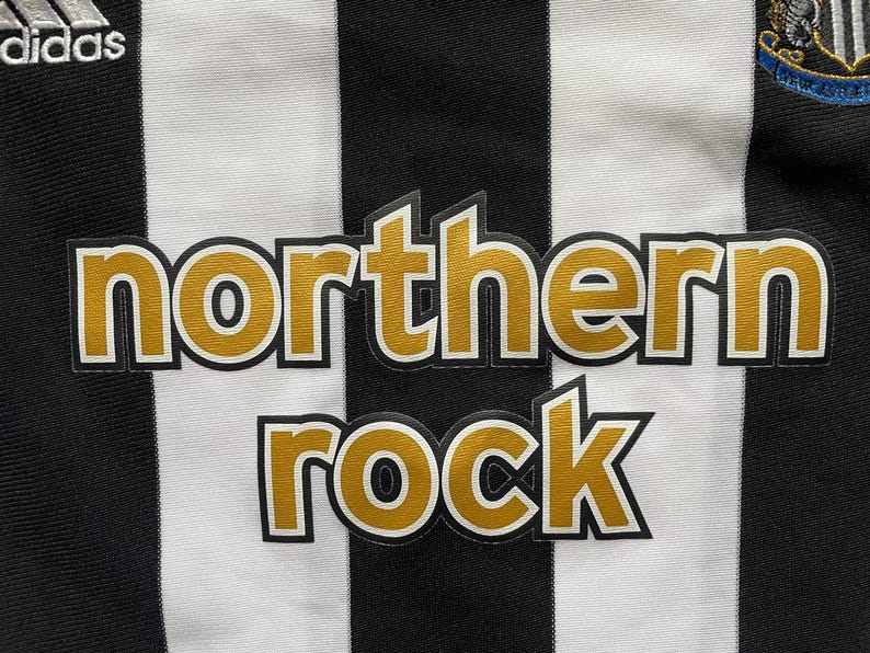 May include: Close-up of a black and white striped jersey. The jersey features the text "northern rock" in gold lettering with a black outline. The Adidas logo is visible in the top left corner.
