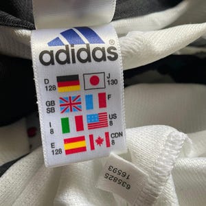 Puede incluir: Primer plano de una etiqueta blanca de Adidas con el logotipo y el texto de la marca. La etiqueta incluye banderas que representan diferentes pa&iacute;ses e informaci&oacute;n de tallas. Tambi&eacute;n es visible una etiqueta m&aacute;s peque&ntilde;a con n&uacute;meros.
