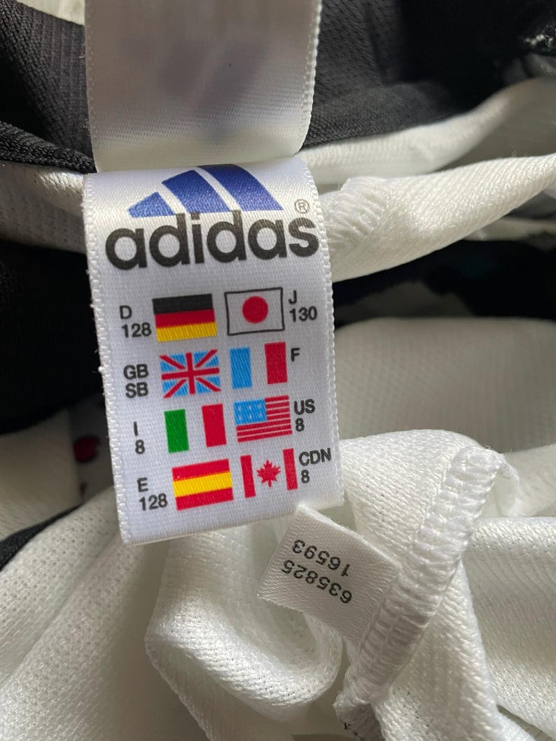 Puede incluir: Primer plano de una etiqueta blanca de Adidas con el logotipo y el texto de la marca. La etiqueta incluye banderas que representan diferentes pa&iacute;ses e informaci&oacute;n de tallas. Tambi&eacute;n es visible una etiqueta m&aacute;s peque&ntilde;a con n&uacute;meros.