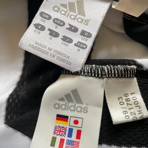 May include: Close-up of two white Adidas labels with black text and logos. One label includes size conversions for UK, USA, E, F, I, J, D, and CDN. The other label displays flags of various countries. The labels are attached to a black and white striped fabric.