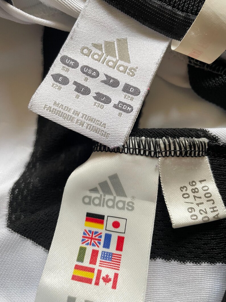 May include: Close-up of two white Adidas labels with black text and logos. One label includes size conversions for UK, USA, E, F, I, J, D, and CDN. The other label displays flags of various countries. The labels are attached to a black and white striped fabric.