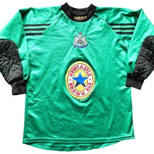 K&ouml;nnte beinhalten: Gr&uuml;nes Langarm-Fu&szlig;balltrikot mit schwarzen Akzenten, das das Wappen von Newcastle United und ein Newcastle Brown Ale-Logo zeigt. Mit schwarzen gepolsterten Ellbogenpolstern und dem Adidas-Logo.