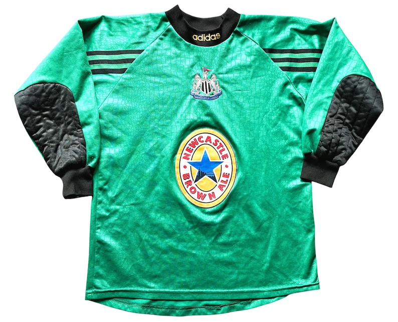 K&ouml;nnte beinhalten: Gr&uuml;nes Langarm-Fu&szlig;balltrikot mit schwarzen Akzenten, das das Wappen von Newcastle United und ein Newcastle Brown Ale-Logo zeigt. Mit schwarzen gepolsterten Ellbogenpolstern und dem Adidas-Logo.