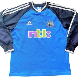 May include: A blue long-sleeved soccer jersey with black and white stripes on the shoulders. The jersey has the Adidas logo, the Newcastle United crest, and the NTL logo.