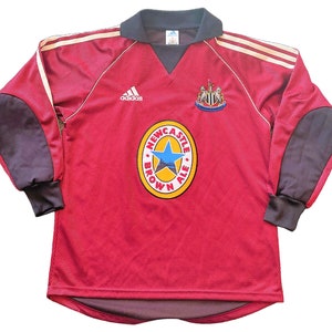 May include: A red long-sleeved football shirt with a dark grey collar and sleeves. It displays the Adidas logo, a team crest, and a "Newcastle Brown Ale" logo. Gold stripes are on the shoulders. The shirt is a vintage design.