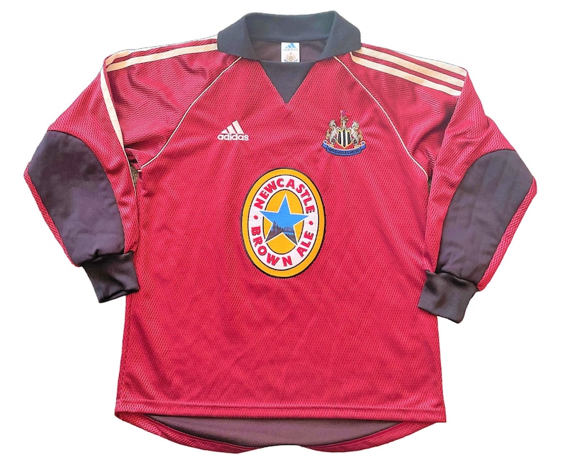 May include: A red long-sleeved football shirt with a dark grey collar and sleeves. It displays the Adidas logo, a team crest, and a "Newcastle Brown Ale" logo. Gold stripes are on the shoulders. The shirt is a vintage design.