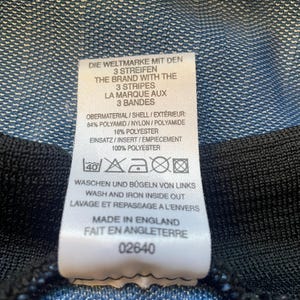 May include: A white tag with black text detailing the care instructions for a garment. The tag reads "Die Weltmarke mit den 3 Streifen", "The Brand with the 3 Stripes", "La Marque aux 3 Bandes", "Obermaterial/Shell/Ext&eacute;rieur: 84% Polyamid/Nylon/Polyamide 16% Polyester", "Einsatz/Insert/Empiecement 100% Polyester", "Waschen und B&uuml;geln von Links", "Wash and Iron Inside Out", "Lavage et Repassage a l'Envers", "Made in England", "Fait en Angleterre", and "02640".