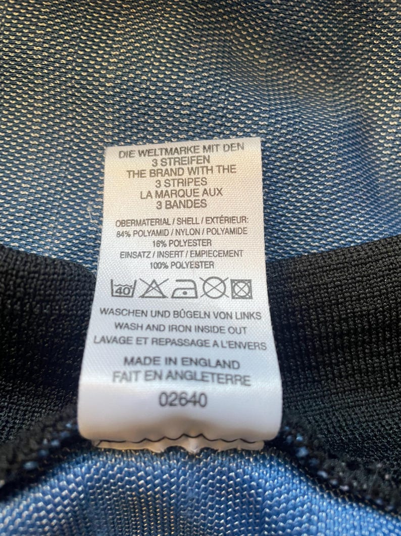 May include: A white tag with black text detailing the care instructions for a garment. The tag reads "Die Weltmarke mit den 3 Streifen", "The Brand with the 3 Stripes", "La Marque aux 3 Bandes", "Obermaterial/Shell/Ext&eacute;rieur: 84% Polyamid/Nylon/Polyamide 16% Polyester", "Einsatz/Insert/Empiecement 100% Polyester", "Waschen und B&uuml;geln von Links", "Wash and Iron Inside Out", "Lavage et Repassage a l'Envers", "Made in England", "Fait en Angleterre", and "02640".