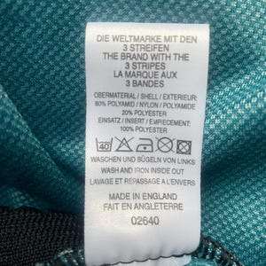 May include: White tag with black text and a washing instruction symbol. The text reads "Die Weltmarke mit den 3 Streifen The Brand with the 3 Stripes La Marque aux 3 Bandes Obermaterial/Shell/Exterieur: 80% Polyamid/Nylon/Polyamide 20% Polyester Einsatz/Insert/Empiecement: 100% Polyester Waschen und Bugeln von Links Wash and Iron Inside Out Lavage et Repassage a l'Envers Made in England Fait en Angleterre 02640"